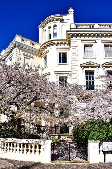 The image depicts a stately white historic building with ornate architectural details, situated behind a garden with blooming cherry blossom trees and neatly maintained greenery. The building features tall windows, decorative cornices, and a rounded corner turret under a clear, bright blue sky. The garden in front is enclosed by a white balustrade and iron gate, with a sign indicating 'Stanhope Crescent.' The scene is well-lit by natural daylight, emphasizing the pristine condition of the building’s façade and the vibrant colors of the flowering trees. This setting exemplifies a well-maintained residence in Notting Hill, highlighting the importance of surface cleaning and upkeep, which is often managed by professional cleaning providers like Notting Hill Carpet Cleaners for both domestic and commercial properties. The overall impression is one of cleanliness and elegance, suitable for a discussion on estate maintenance or deep cleaning services.