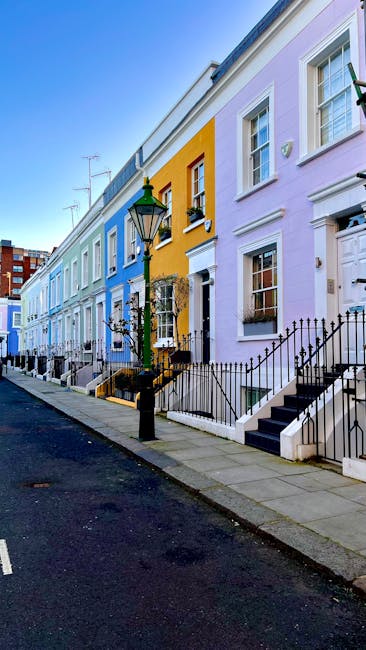 A row of colorful terraced houses in Notting Hill, featuring facades painted in pastel shades of blue, yellow, pink, and lavender. Each house has a small front porch with steps leading up to white doors, black wrought-iron railings, and potted plants near the entrances. The street scene is illuminated by a traditional black street lamp, and the sky is clear with hints of sunlight. Notting Hill Carpet Cleaners specializes in surface cleaning and deep cleaning services for residential properties in this area, ensuring homes are hygienic and well-maintained.