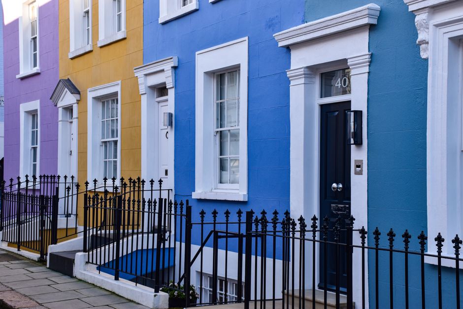 Exterior view of a row of Victorian terraced houses in Notting Hill, featuring pastel-colored facades with decorative columns painted in shades of teal, blue, and pink. The houses have large sash wind