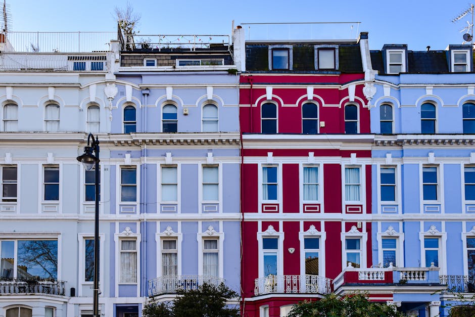 A row of three Victorian-style residential buildings with ornate architectural details, painted in pastel shades of lavender, pink, and blue. The facades feature decorative moldings, tall narrow windows with white frames, and balconies with intricate white iron railings. A black street lamp is mounted on a post in front of the buildings, and the sky is clear with bright sunlight illuminating the surfaces. The image reflects well-maintained exteriors, demonstrating effective surface cleaning and preservation by Notting Hill Carpet Cleaners as part of their domestic cleaning services.