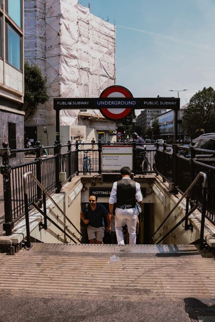 This image depicts an urban underground entrance to the Notting Hill Gate station with black metal railings and concrete steps leading down. Two men are seen descending, one dressed in dark clothing wearing sunglasses, and the other in a white shirt with a dark vest. Above, a large underground sign displays the London Underground logo with the words 'Public Subway' and 'Underground' on a black background. In the background, a building covered with white scaffolding and fabric wraps is visible, indicating construction or maintenance work. The scene is illuminated by natural daylight, with a clear sky overhead, and a few streetlights and vehicles parked nearby. This setting emphasizes the typical city infrastructure around public transit hubs, fitting within a context where professional cleaning or maintenance services, such as those provided by Notting Hill Carpet Cleaners, might be relevant to maintaining cleanliness in high-traffic urban areas.